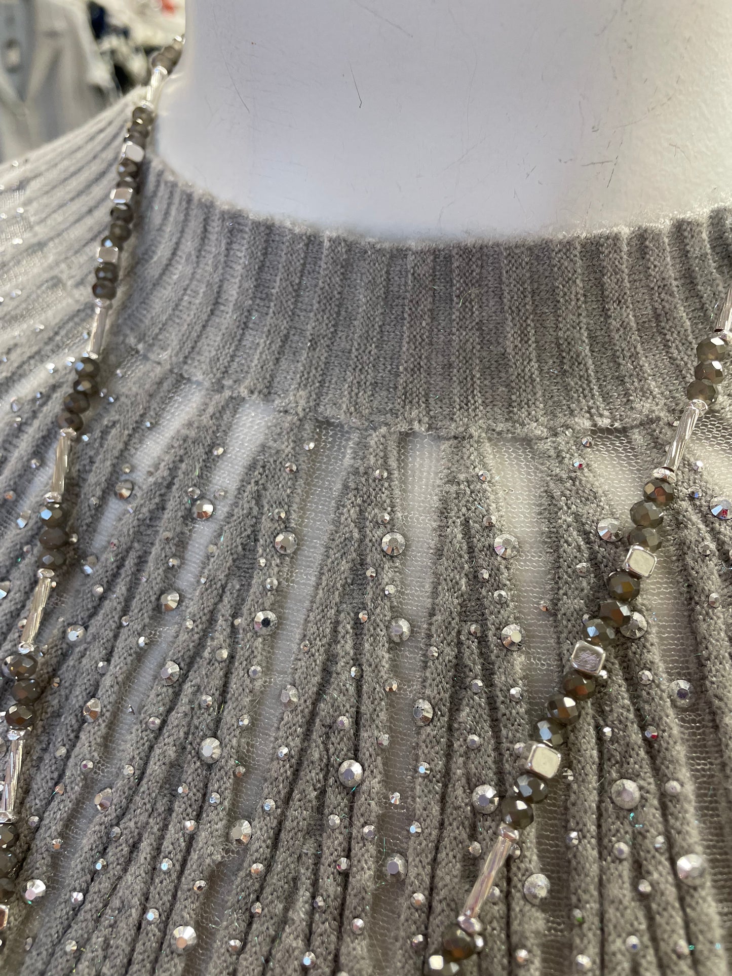 A mannequin in a retail store showcases the Nu-Look Fashions High Neck L/S Top, a light gray ribbed long-sleeve sweater with gem details, accessorized with a long silver necklace.