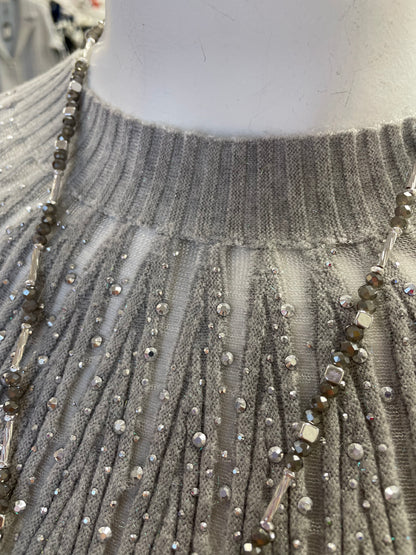 A mannequin in a retail store showcases the Nu-Look Fashions High Neck L/S Top, a light gray ribbed long-sleeve sweater with gem details, accessorized with a long silver necklace.