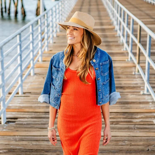 A woman in the Wallaroo Klara Wide Brim Sun Hat, orange dress, and denim jacket walks on a wooden pier with water to the left, showcasing stylish sun protection.