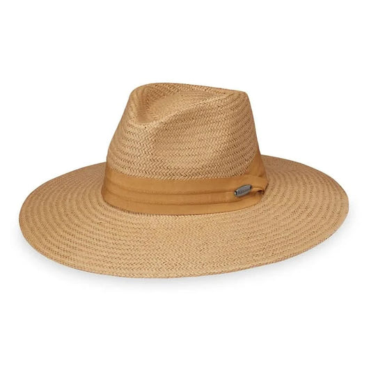 A woman in the Wallaroo Klara Wide Brim Sun Hat, orange dress, and denim jacket walks on a wooden pier with water to the left, showcasing stylish sun protection.