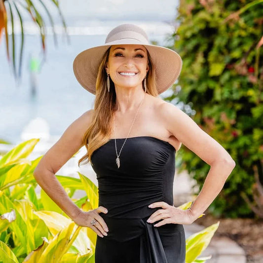 A woman in a strapless black dress smiles outdoors, wearing the Wallaroo Margot Wide Brim Sun Hat, with greenery and water in the background.