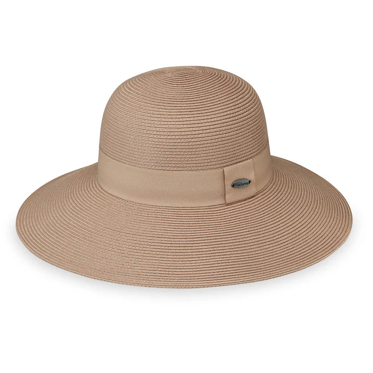 A woman in a strapless black dress smiles outdoors, wearing the Wallaroo Margot Wide Brim Sun Hat, with greenery and water in the background.