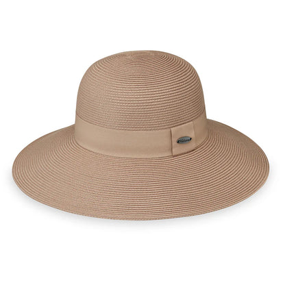A woman in a strapless black dress smiles outdoors, wearing the Wallaroo Margot Wide Brim Sun Hat, with greenery and water in the background.