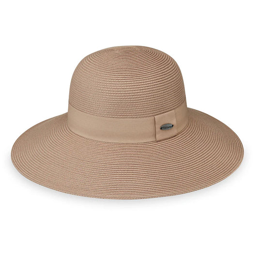 A woman in a strapless black dress smiles outdoors, wearing the Wallaroo Margot Wide Brim Sun Hat, with greenery and water in the background.