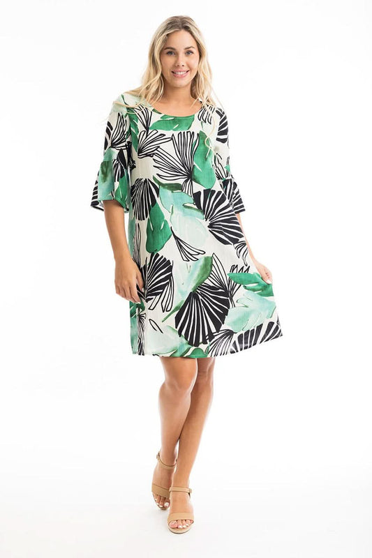 A woman models the Orientique Papagayo Easy Fit Frill Dress, a loose, knee-length piece with a green and black tropical leaf print and beige heeled sandals, perfect for summer, against a white background.