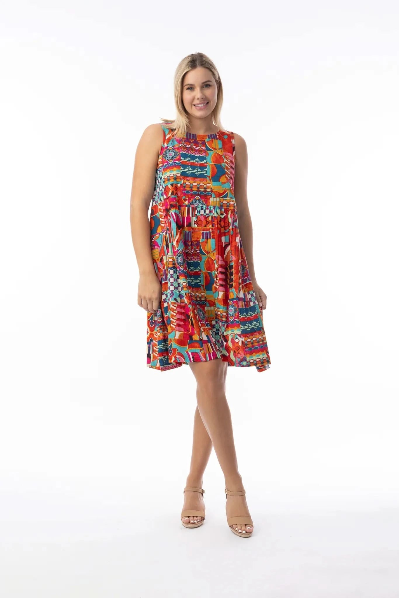 A woman stands on a white background wearing the Orientique Peru Print Dress, a sleeveless, knee-length dress with a colorful retro geometric pattern, paired with tan heeled sandals.