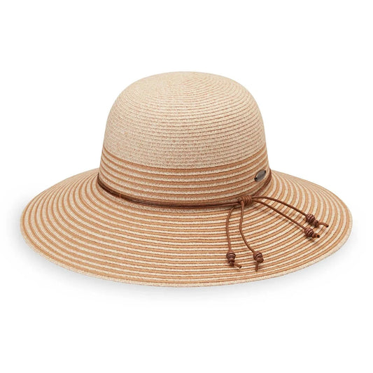 A woman smiles indoors, wearing the Wallaroo Petite Marseille wide-brim straw hat made from Flexi-Weave fabric with an adjustable fit and an orange off-the-shoulder top, holding the brim of her hat.