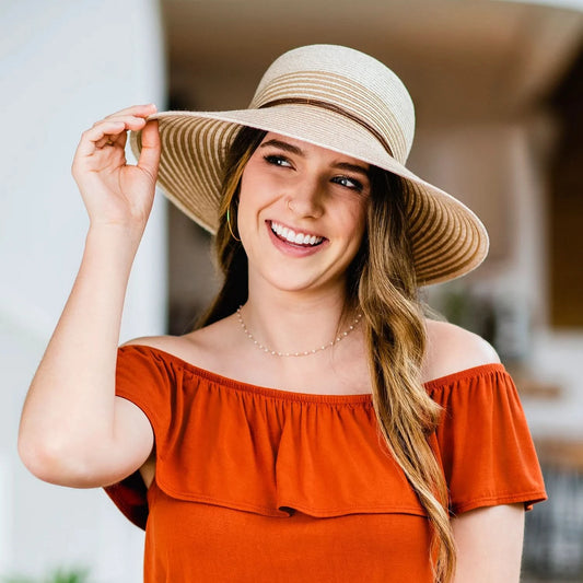 A woman smiles indoors, wearing the Wallaroo Petite Marseille wide-brim straw hat made from Flexi-Weave fabric with an adjustable fit and an orange off-the-shoulder top, holding the brim of her hat.