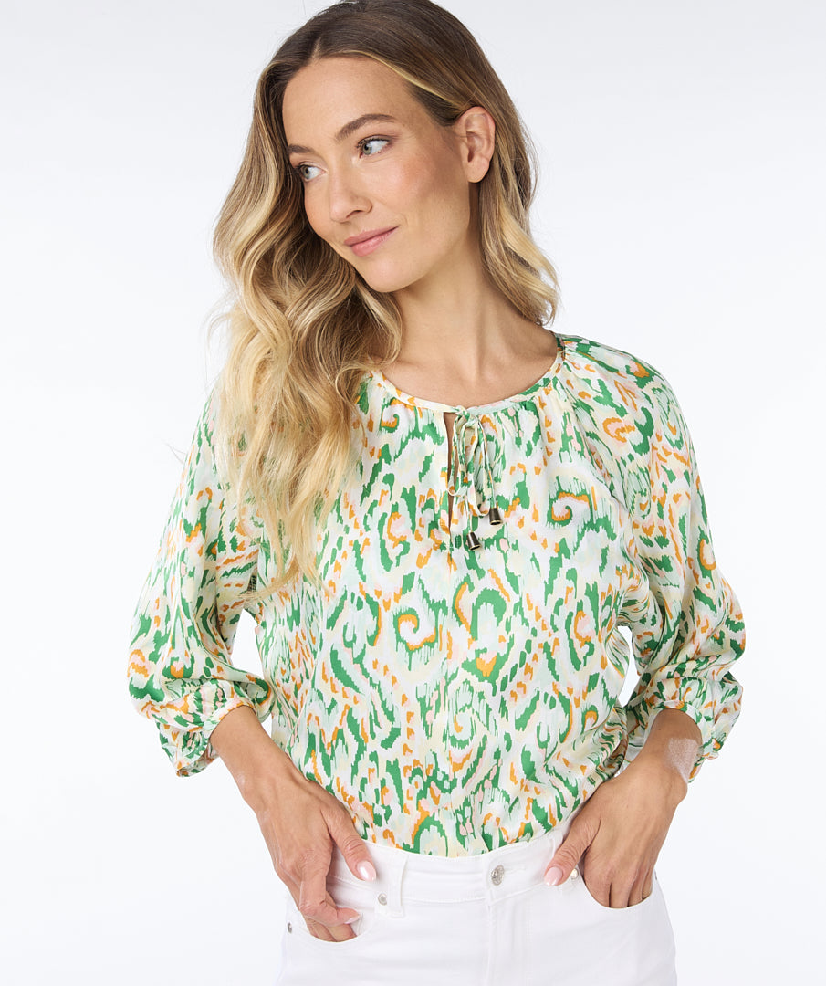A person wearing an Esqualo Pastel Blouse and white pants stands against a white background, gently touching their hair.