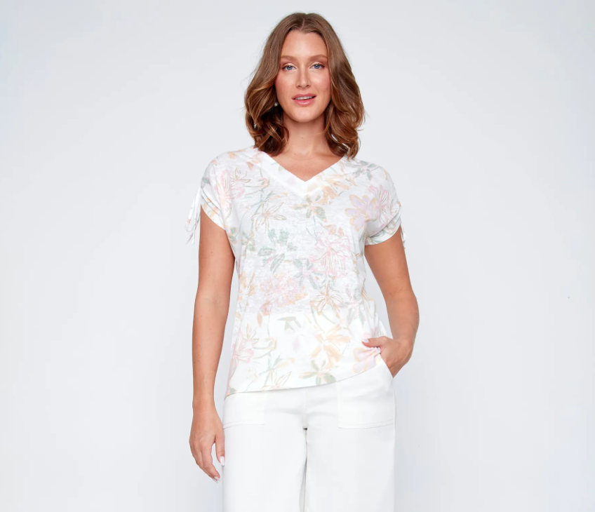 A woman models the CoCo Y Club V-neck top, a light floral, short-sleeve blouse, paired with white pants. The perfect spring-summer choice for effortless style.