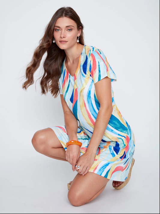 A woman with long brown hair kneels on one knee, wearing CoCo Y Club's Mini Flow Dress—a vibrant, short-sleeved summer dress with an abstract print—paired with earrings and an orange bangle.