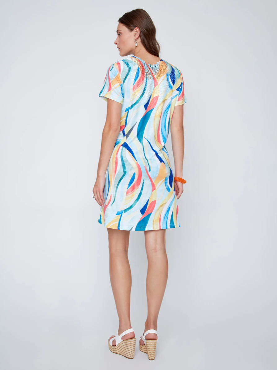 A woman with long brown hair kneels on one knee, wearing CoCo Y Club's Mini Flow Dress—a vibrant, short-sleeved summer dress with an abstract print—paired with earrings and an orange bangle.