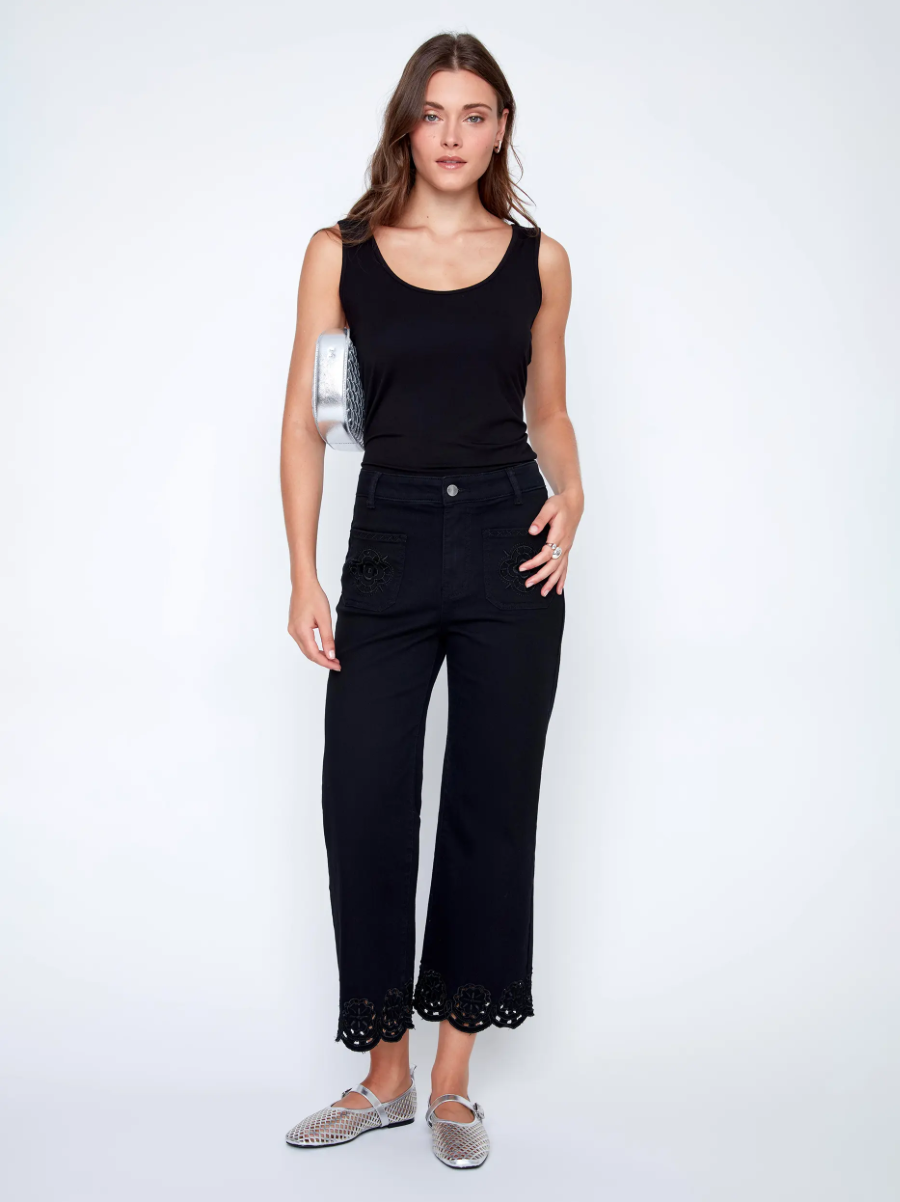 A woman in a black sleeveless top and CoCo Y Club Embroidered Black Trousers holds a silver clutch and wears silver slip-on shoes, standing against a plain white background.