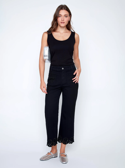 A woman in a black sleeveless top and CoCo Y Club Embroidered Black Trousers holds a silver clutch and wears silver slip-on shoes, standing against a plain white background.