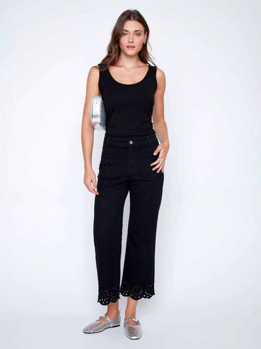 A woman in a black sleeveless top and CoCo Y Club Embroidered Black Trousers holds a silver clutch and wears silver slip-on shoes, standing against a plain white background.