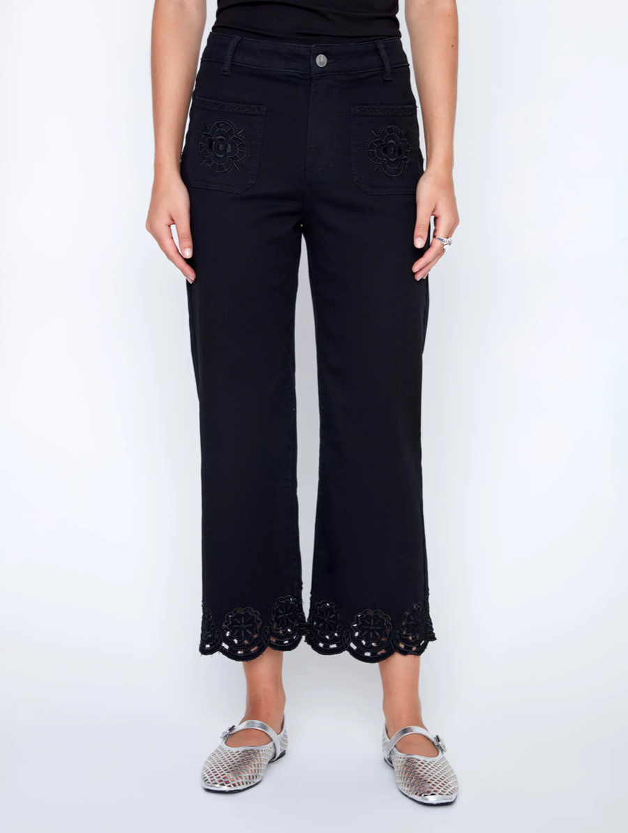 A woman in a black sleeveless top and CoCo Y Club Embroidered Black Trousers holds a silver clutch and wears silver slip-on shoes, standing against a plain white background.