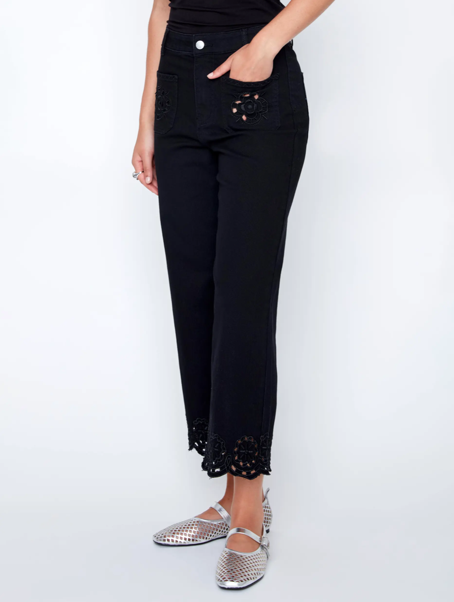 A woman in a black sleeveless top and CoCo Y Club Embroidered Black Trousers holds a silver clutch and wears silver slip-on shoes, standing against a plain white background.