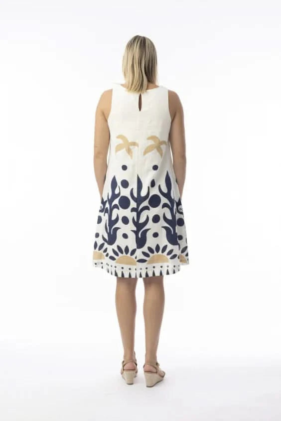 A woman in an Orientique Nazca Sleeveless Midi Dress with bold blue and tan abstract patterns and a flared hem stands by a black iron fence on a sunny day.