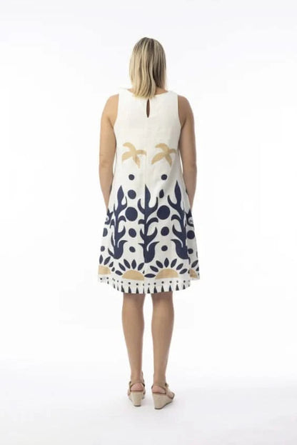 A woman in an Orientique Nazca Sleeveless Midi Dress with bold blue and tan abstract patterns and a flared hem stands by a black iron fence on a sunny day.