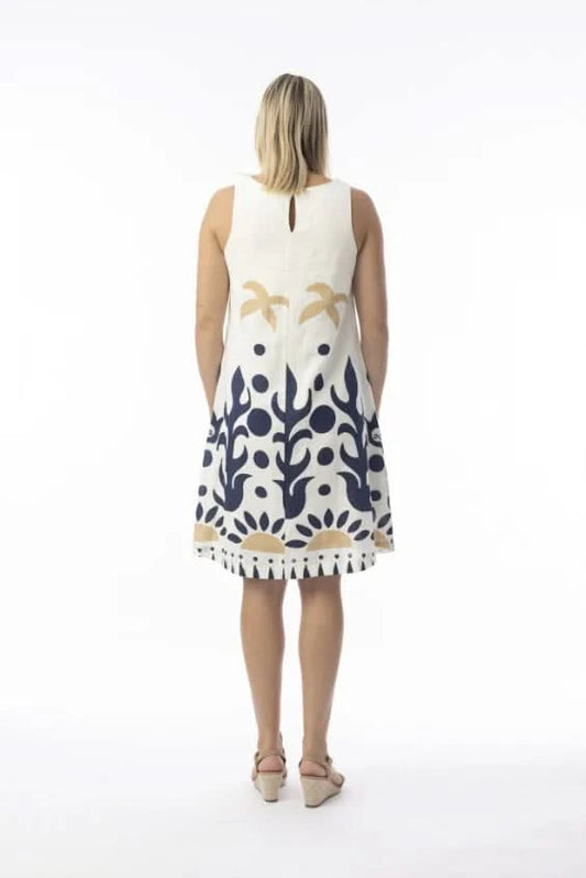 A woman in an Orientique Nazca Sleeveless Midi Dress with bold blue and tan abstract patterns and a flared hem stands by a black iron fence on a sunny day.