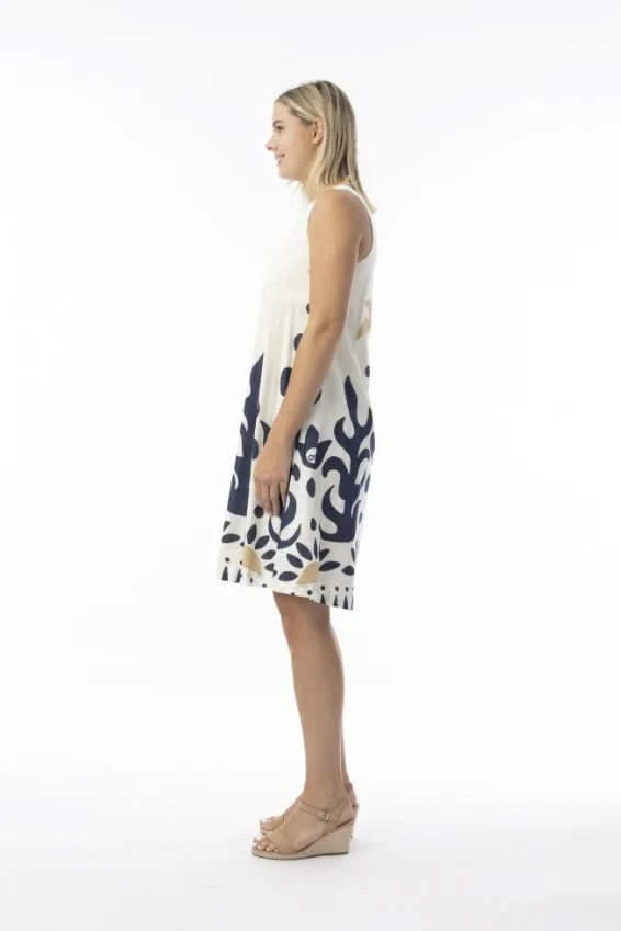 A woman in an Orientique Nazca Sleeveless Midi Dress with bold blue and tan abstract patterns and a flared hem stands by a black iron fence on a sunny day.