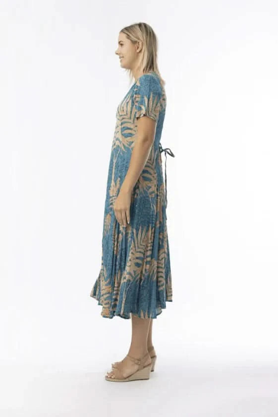 A woman stands in profile against a white background, wearing the Orientique Ponta Verde Short Sleeve Godet Dress in blue and tan floral, paired with tan wedge sandals—an effortless addition to any summer wardrobe.