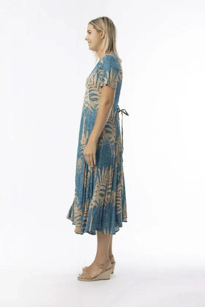 A woman stands in profile against a white background, wearing the Orientique Ponta Verde Short Sleeve Godet Dress in blue and tan floral, paired with tan wedge sandals—an effortless addition to any summer wardrobe.