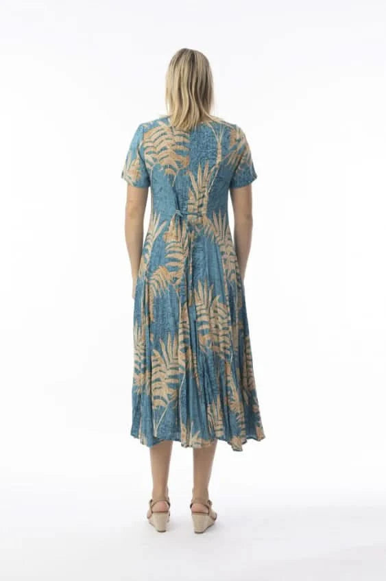 A woman stands in profile against a white background, wearing the Orientique Ponta Verde Short Sleeve Godet Dress in blue and tan floral, paired with tan wedge sandals—an effortless addition to any summer wardrobe.