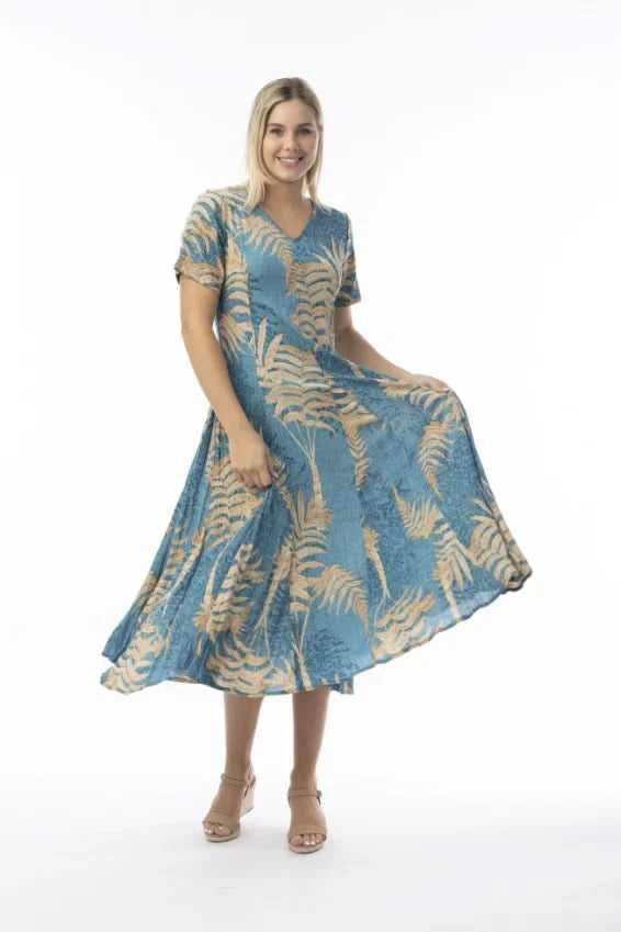 A woman stands in profile against a white background, wearing the Orientique Ponta Verde Short Sleeve Godet Dress in blue and tan floral, paired with tan wedge sandals—an effortless addition to any summer wardrobe.