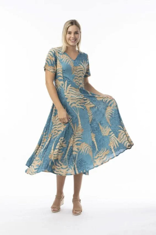 A woman stands in profile against a white background, wearing the Orientique Ponta Verde Short Sleeve Godet Dress in blue and tan floral, paired with tan wedge sandals—an effortless addition to any summer wardrobe.