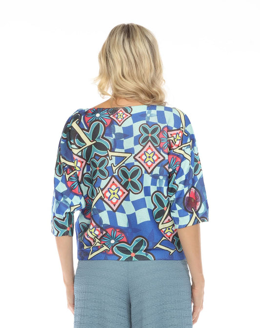 A person with shoulder-length blonde hair is shown from behind, wearing the Inoah 3/4 Sleeve Dolman Sweater featuring a colorful geometric and floral pattern, paired with blue textured pants.