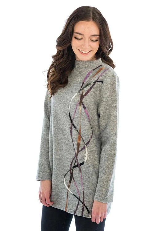 A woman wearing the Trisha Tyler Side Felted Funnel Neck Tunic, designed with multicolored abstract lines on a gray background, stands against a white backdrop, looking down and smiling.
