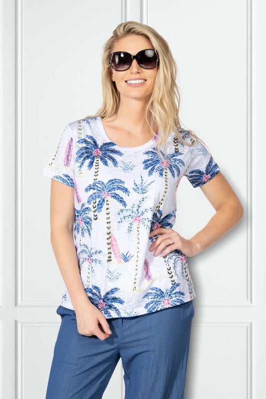 A woman wearing sunglasses and blue pants smiles in front of a white panel wall, modeling the Variations Short Sleeve Top with a blue and pink tropical palm tree print.
