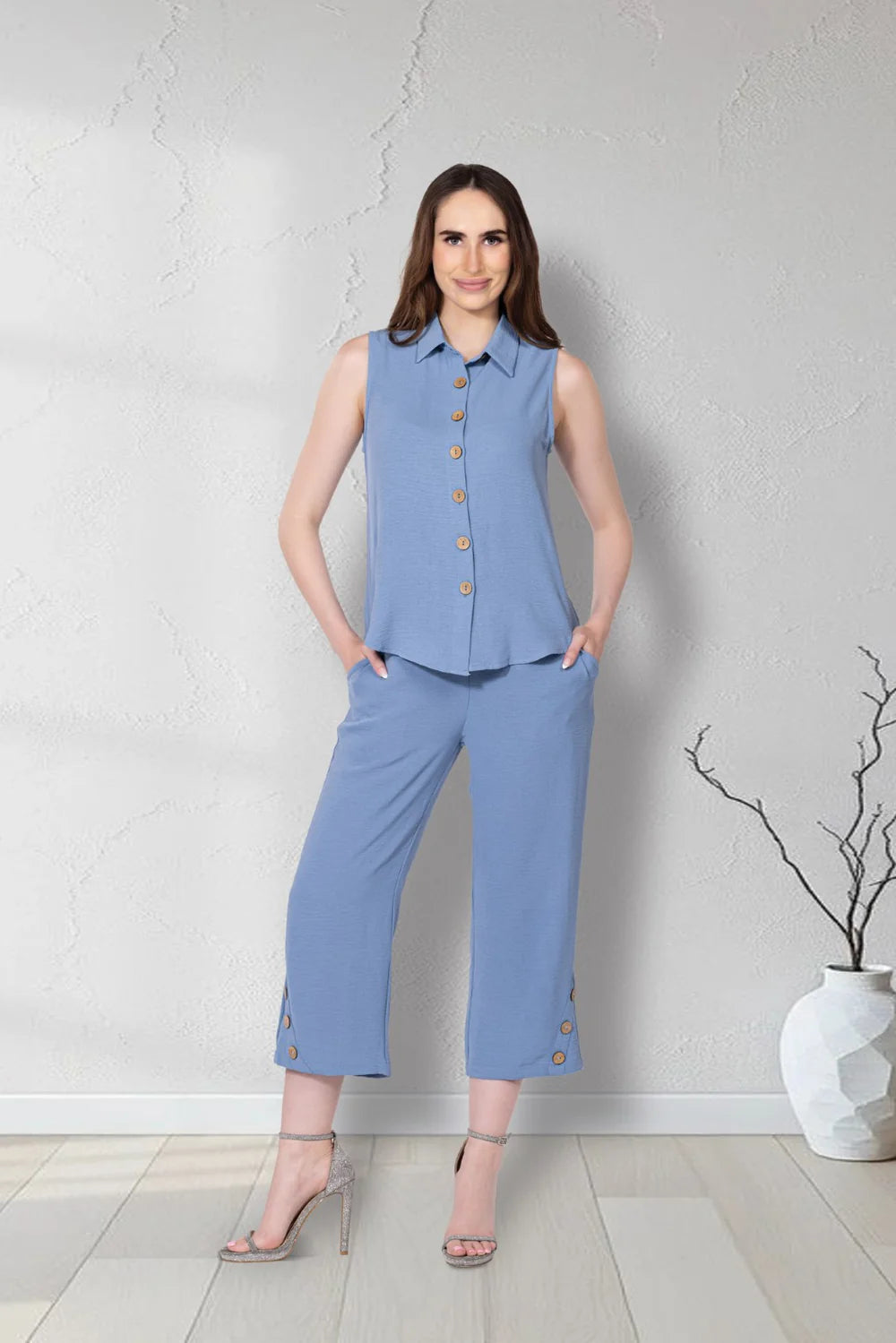 A woman models Variations' Button-Hem Capri Pants and a sleeveless steel blue collared top with gold button accents, standing indoors on a light wood floor near a vase with bare branches.