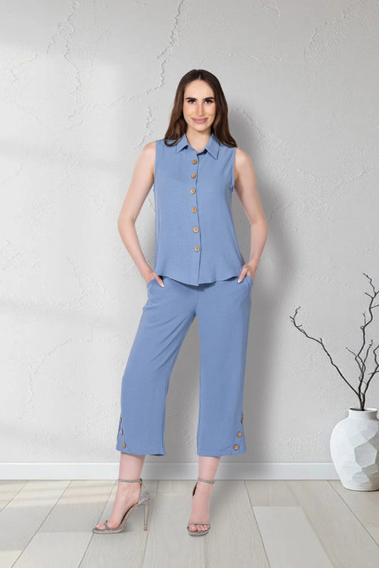A woman models Variations' Button-Hem Capri Pants and a sleeveless steel blue collared top with gold button accents, standing indoors on a light wood floor near a vase with bare branches.