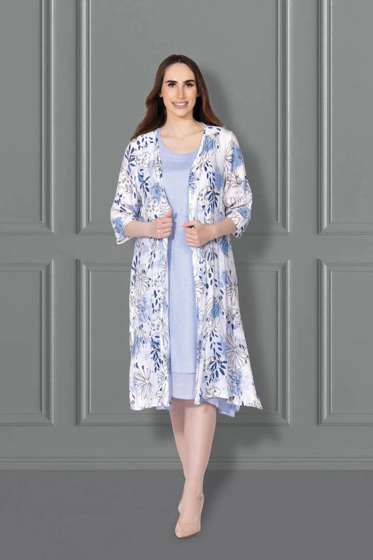 A woman stands against a gray paneled wall, wearing the Variations Solid Dress with Floral Print Jacket Set—a chic two-piece featuring a light blue dress and elegant floral overcoat, paired with beige heels. Perfect for stylish occasions.