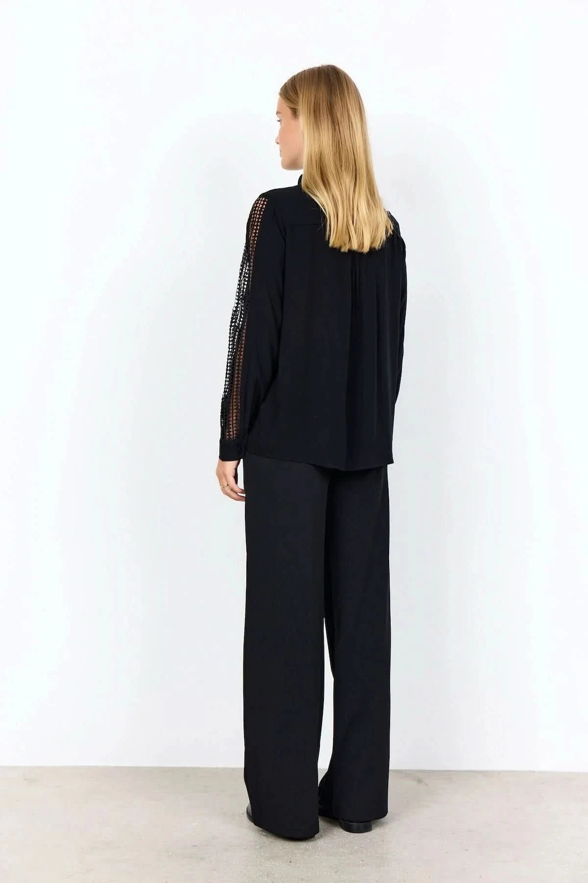 A woman stands against a plain background wearing the Soya Concepts Ladies Woven Blouse—a black, lightweight viscose blouse with lace long sleeves—paired with black pants.