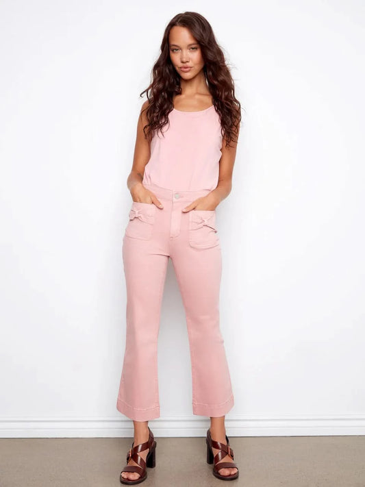 A woman stands against a white wall wearing Charlie B's Flare Twill Pants With Bow Patch Pockets, a sleeveless pink top, and brown platform sandals, with her hands in her front pockets.