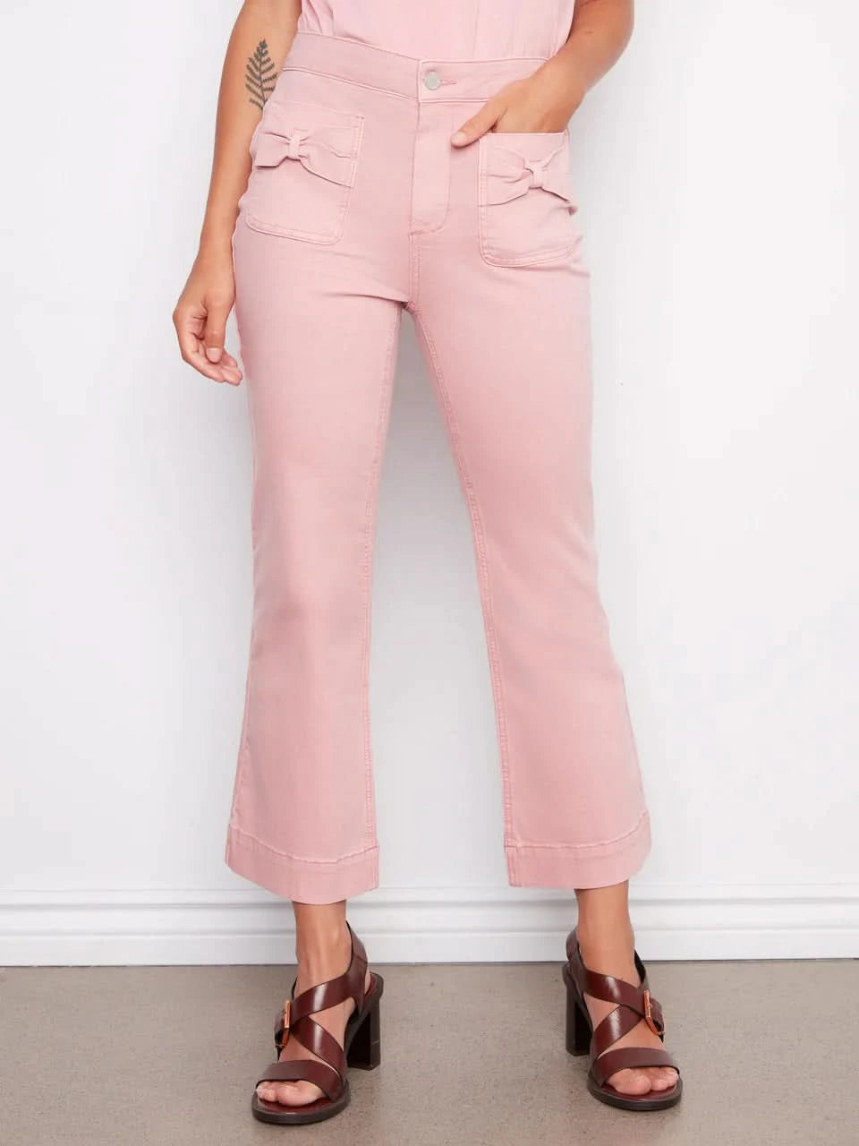 A woman stands against a white wall wearing Charlie B's Flare Twill Pants With Bow Patch Pockets, a sleeveless pink top, and brown platform sandals, with her hands in her front pockets.