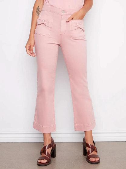 A woman stands against a white wall wearing Charlie B's Flare Twill Pants With Bow Patch Pockets, a sleeveless pink top, and brown platform sandals, with her hands in her front pockets.