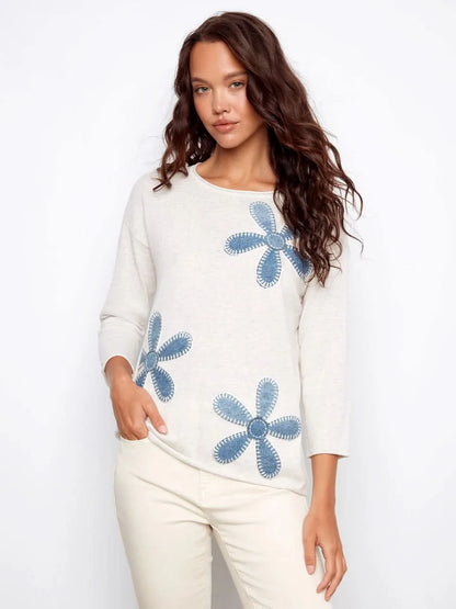 A woman wearing the Charlie B Floral Denim Patch Sweater, a light long-sleeve top with blue floral details, paired with light pants and standing against a plain background.