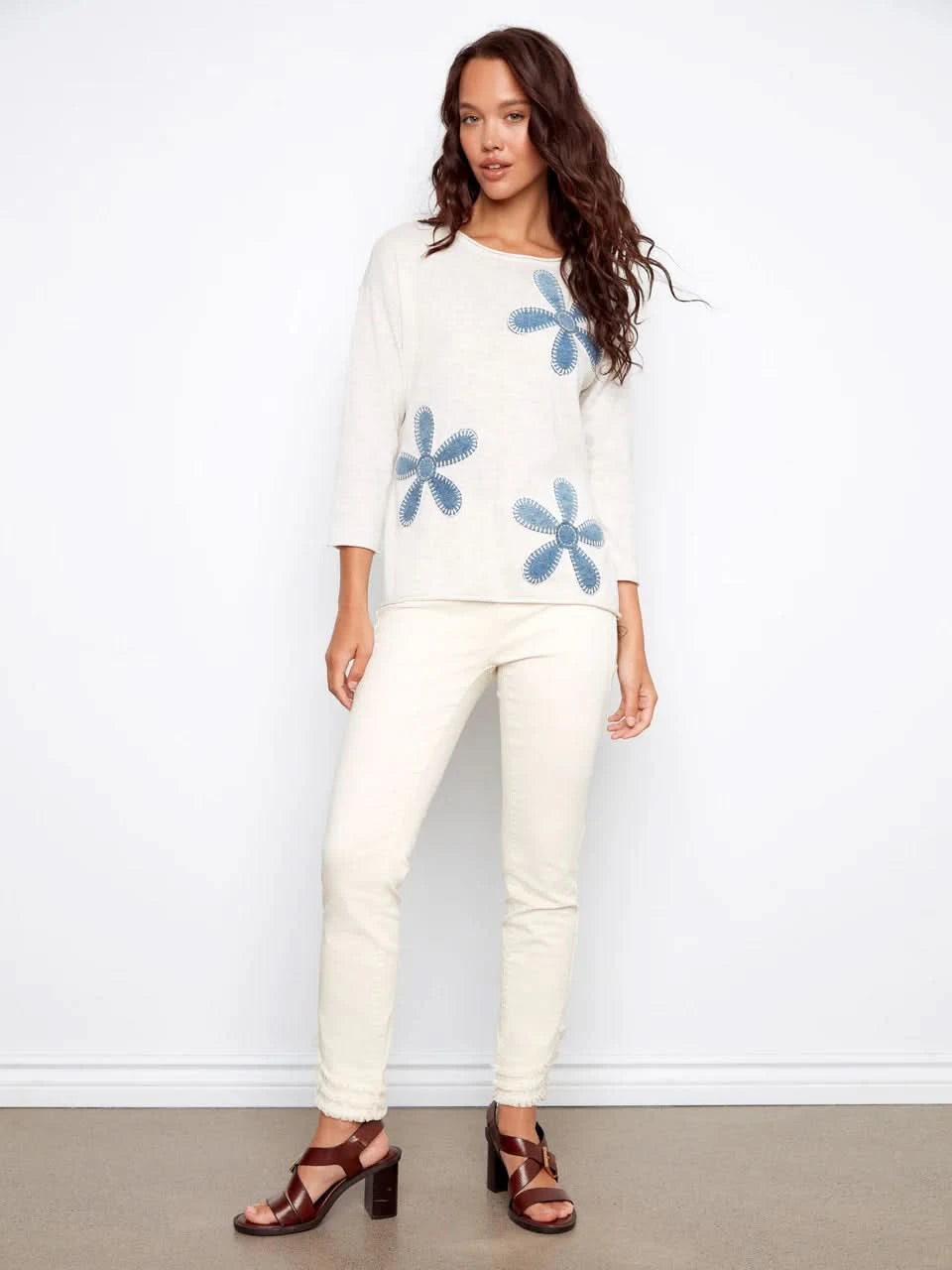 A woman wearing the Charlie B Floral Denim Patch Sweater, a light long-sleeve top with blue floral details, paired with light pants and standing against a plain background.