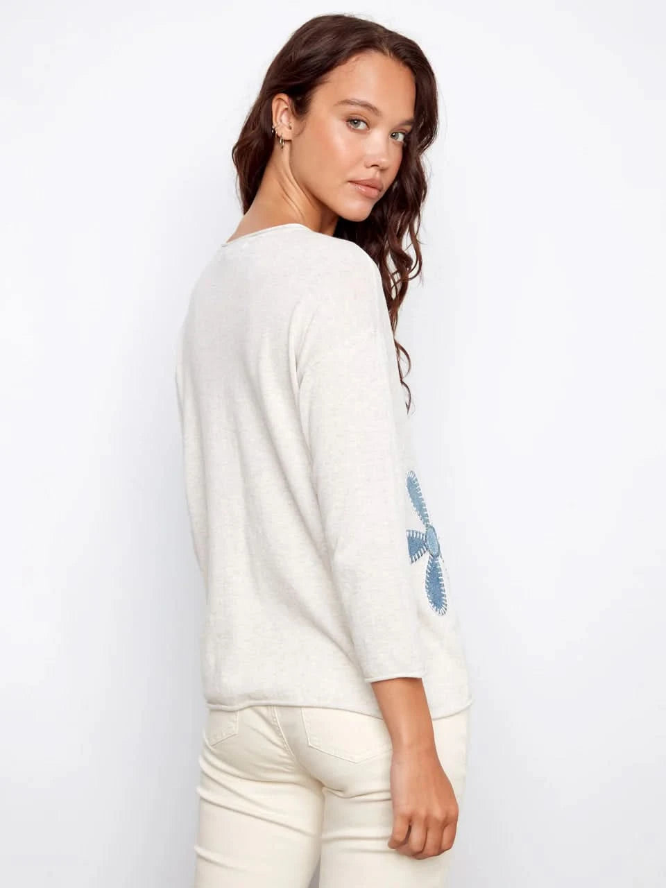 A woman wearing the Charlie B Floral Denim Patch Sweater, a light long-sleeve top with blue floral details, paired with light pants and standing against a plain background.