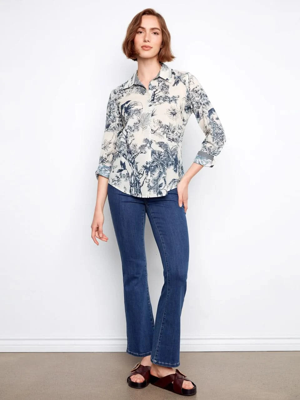 A woman wears a Charlie B Printed Cotton Voile Button-Up Blouse with a blue and white botanical pattern, paired with blue jeans, standing against a plain light background.