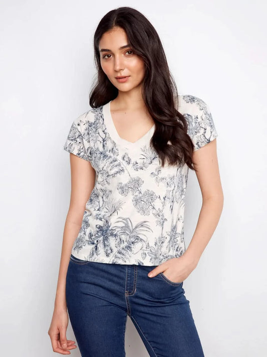A woman models the Charlie B Printed Organic Cotton Drop Shoulder V-Neck T-Shirt - Versailles with blue jeans, posing against a plain white background.