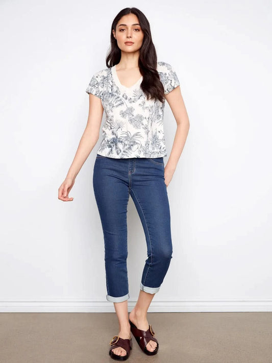 A woman models the Charlie B Printed Organic Cotton Drop Shoulder V-Neck T-Shirt - Versailles with blue jeans, posing against a plain white background.