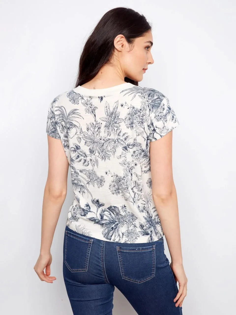A woman models the Charlie B Printed Organic Cotton Drop Shoulder V-Neck T-Shirt - Versailles with blue jeans, posing against a plain white background.