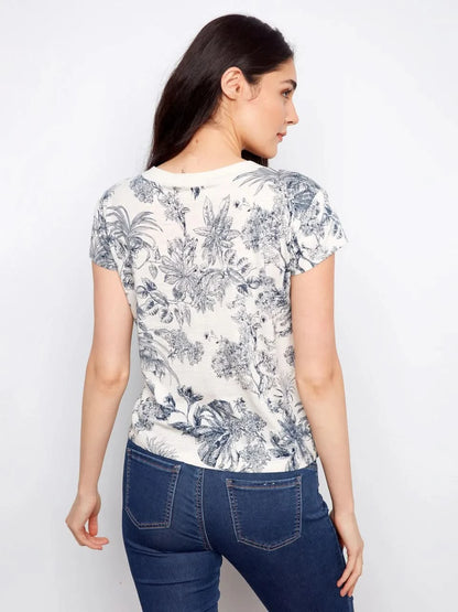 A woman models the Charlie B Printed Organic Cotton Drop Shoulder V-Neck T-Shirt - Versailles with blue jeans, posing against a plain white background.