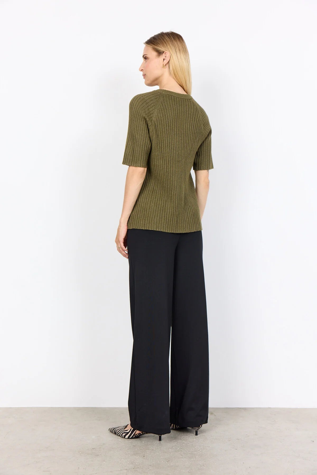 A person wearing the Soya Concepts Dark Olive Long Sleeved Knit in a relaxed fit with black pants stands against a plain white background, hands in pockets.