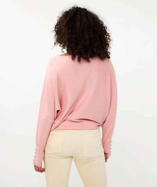 A person with curly hair is standing with their back to the camera, wearing Esqualo's pink Batwing Sweater with buttoned cuffs and light-colored pants.